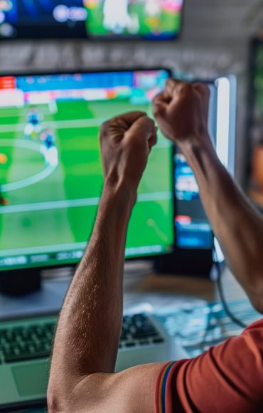 A man clenches his fist in victory in front of a computer screen displaying a live soccer game, reflecting the thrill of potentially winning in online betting.